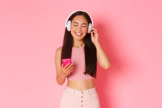 Portrait of carefree attractive asian woman enjoying favorite song, close eyes to relax while listening music in hearphones, holding mobile phone and standing over pink background