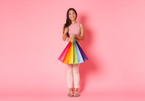 Full length portrait of beautiful and trendy asian girl shopaholic, holding shopping bags and pointing at herself, bought summer clothes for her vacation, standing pink background