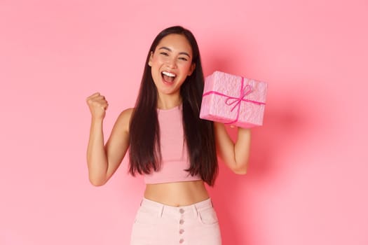 Holidays, celebration and lifestyle concept. Triumphing happy asian cute birthday girl looking upbeat, likes receiving gifts, raising fist pump and showing wrapped present, standing pink background.