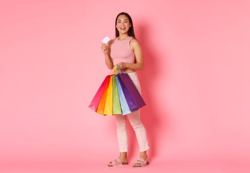 Full length portrait of upbeat and excited, smiling trendy asian girl showing her credit card and looking pleased, carry shopping bags, bought new clothes for vacation, pink background