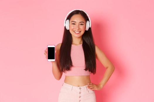 Technology, communication and online lifestyle concept. Smiling beautiful asian girl promote mobile phone application, showing smartphone screen while listening music in headphones