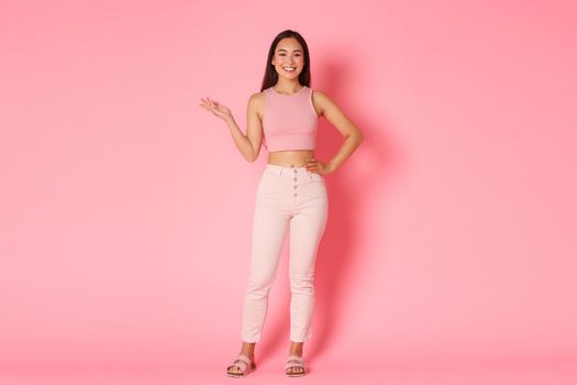 Fashion, beauty and lifestyle concept. Full-length portrait of attractive tall asian girl smiling and holding left arm raised, demonstrating product over pink background