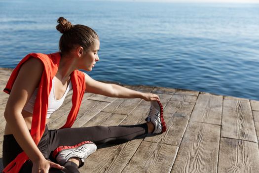 Outdoor shot of attractive fitness woman warming-up before jogging, sitting on pier and stretching legs