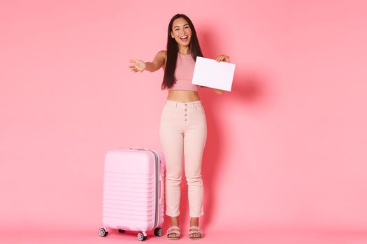 Travelling, holidays and vacation concept. Full-length of friendly asian female meeting person in airport, standing with suitcase and piece of paper, extend hand for welcome hug, pink background