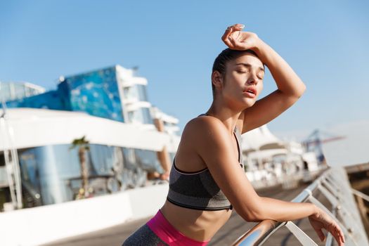 Close-up of attractive young fitness woman leaning on handrail, wiping sweat off forehead after workout, finish jogging on the seaside promenade