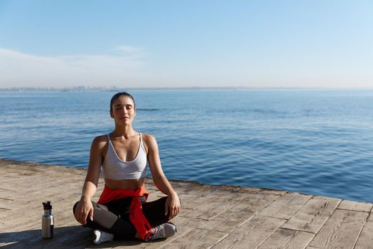 Outdoor shot of peaceful fitness woman sitting in asana and doing yoga near the sea. Female athlete meditating on seaside