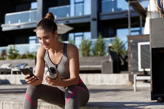 Outdoor shot of attractive sportswoman sitting on bench and using mobile phone, holding water bottle