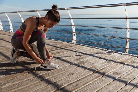 Young attractive fitness woman tying her shoes, jogging on the seaside promenade