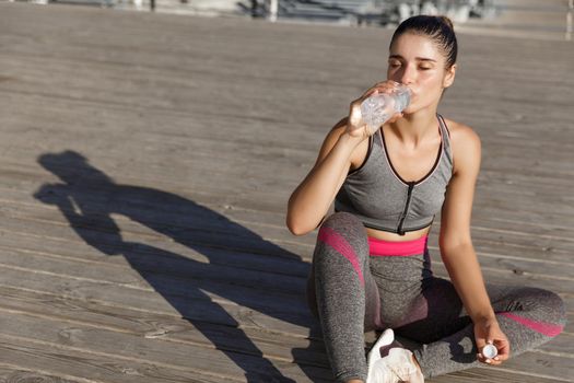 Close-up of attractive sportswoman taking a break after workout, sitting at the seaside and drinking water, looking pleased