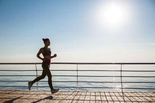 Image of attractive sportswoman jogging alone near the sea. Fitness woman running along seaside