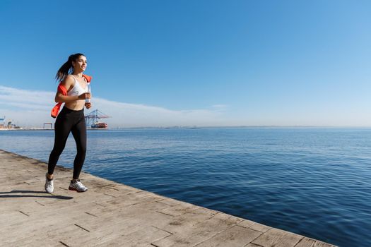 Outdoor shot of happy sportswoman jogging in the morning on a pier, running forward and smiling