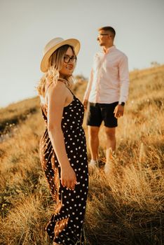 young man and woman walking in the meadow at sunset in summer near the lake. A couple of fair-haired fair-skinned people in love are resting in nature in a field at sunset.