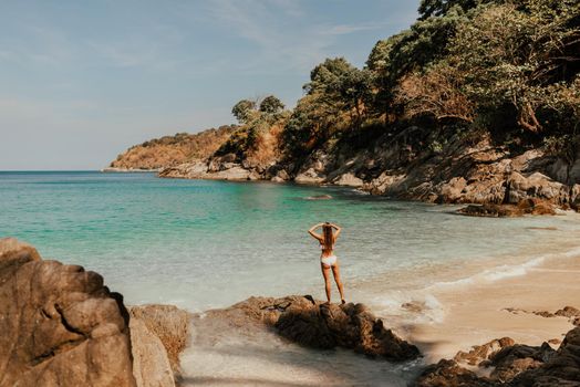 European woman in bikini swimsuit stands backwards on stone rocks on ocean shore