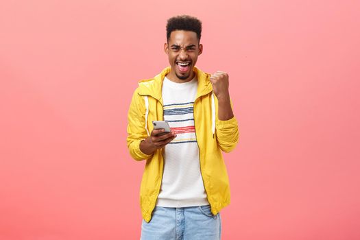 Yes I won. Portrait of happy excited and satisfied african american male with beard raising fist in victory and triumph gesture rejoicing holding smartphone winning in online phone game over pink wall