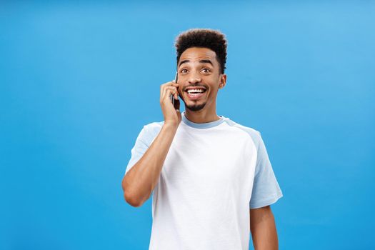 Friendly joyful carefree dark-skinned guy with beard in casual t-shirt talking happily about amusing things holding smartphone near ear having funny, amusing conversation over blue wall