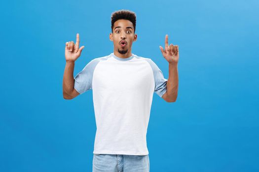 Impressed thrilled handsome african american boyfriend in casual t-shirt folding lips in wow sound being amazed and surprised with cool product pointing up with raised hands over blue wall