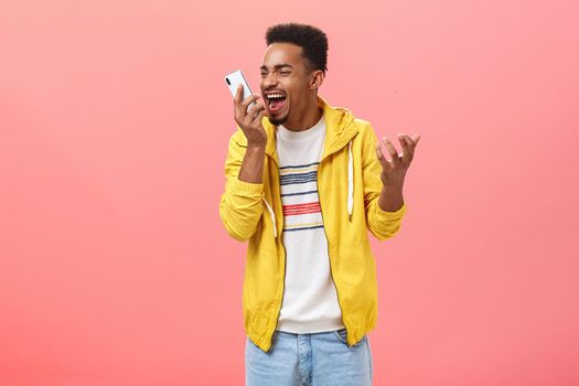 Guy being fed up with dumb customer support shouting at smartphone screen yelling from pissed outrage feelings clenching gist holding cellphone near face over pink background