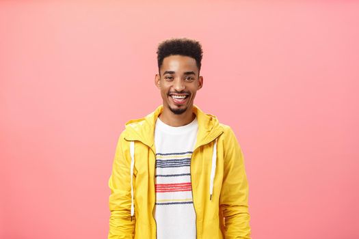 Waist-up shot of charming friendly good-looking dark-skinned male with beard and afro hairstyle in trendy yellow jacket smiling with satisfied pleased grin standing over pink background