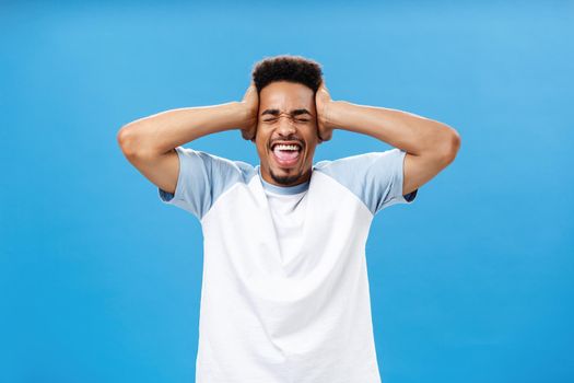 Mind blowing out from lots of information. Portrait of distressed African American male student in t-shirt holding hands on head and yelling with closed eyes losing temper distressed over blue wall