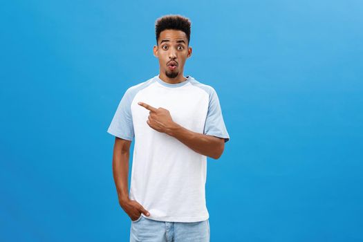 Impressed curious handsome young male with beard saying wow raising eyebrows in wonder pointing left astonished and excited standing thrilled in t-shirt over blue background