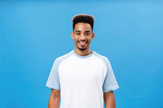 Charming friendly and polite dark-skinned guy with beard and afro hairstyle in trendy t-shirt smiling broadly gazing at camera full of hopes, energetic to start own business over blue wall