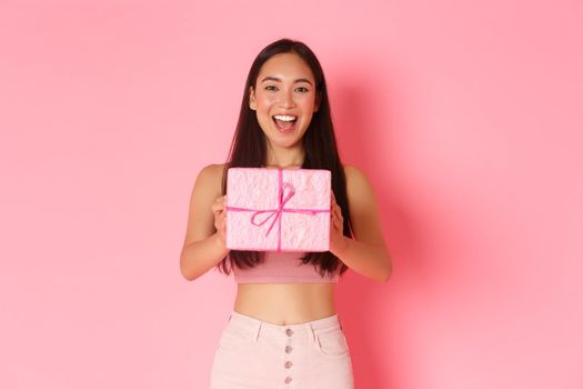 Holidays, celebration and lifestyle concept. Waist-up of cheerful beautiful asian girl making gift, giving friend birthday present in wrapped box, standing pink background, congratulate.