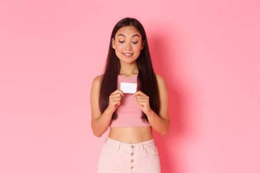 Beauty, fashion and lifestyle concept. Portrait of excited attractive asian girl in trendy clothes tempting to use credit card and buy something, likes shopping, standing pink background
