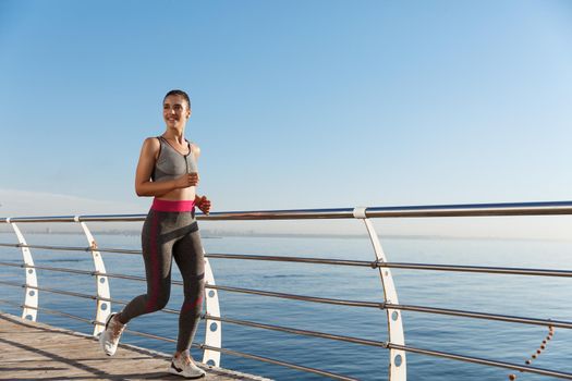Image of happy fitness woman jogging along seaside promenade, smiling during workout