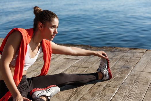 Close-up of confident sportswoman stretching legs, sitting on pier and training