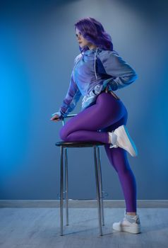a girl in stylish purple sportswear and with purple hair poses sexually on a bar stool