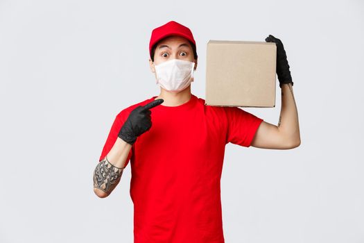 Concept of delivery and carriers during coronavirus pandemic. Wonder what inside. Curious asian courier holding box on shoulder and pointing at package, wear protective gloves and medical mask