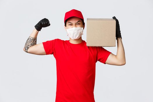 Concept of delivery during coronavirus pandemic. Courier show-off his strength, flex biceps and carry heavy package on shoulder, wear medical mask and gloves for safety on covid-19 quarantine