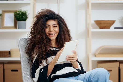 Happy smiling pretty laughing curly tanned Latin woman in striped shirt hold notebook look at camera write work letter essay in home interior. Copy space Banner. Writer concept