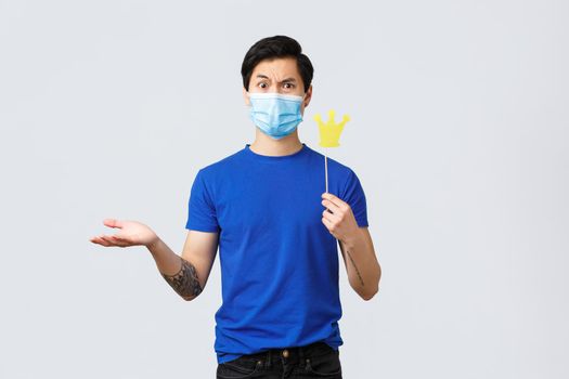 Lifestyle, people different emotions and covid-19 pandemic concept. Confused young arrogant asian guy in medical mask holding crown on stick and shrugging, cant understand