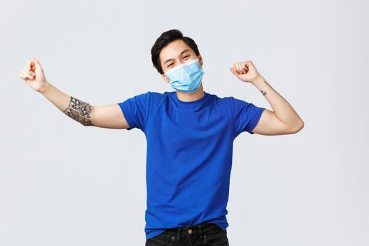 Different emotions, social distancing, self-quarantine on covid-19 concept. Carefree happy asian man celebrating good news with victory dance, feeling excited, hear good news, wear medical mask