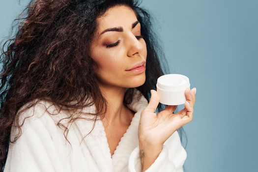 Pensive curly Latin lady in bathrobe Show opened jar of cream Recommend beauty product looking down posing isolated on pastel blue background. Cosmetic product ad concept. Cool offer