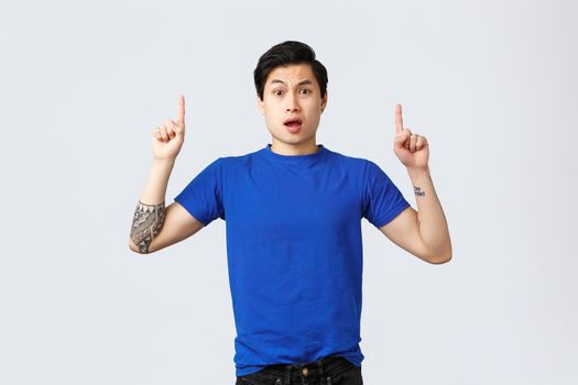 Different emotions, people lifestyle and advertising concept. Concerned and frustrated young asian man cant understand what happened, look confused and pointing fingers up, grey background