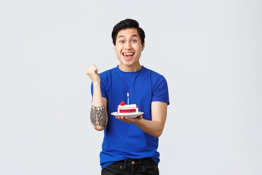 Self-quarantine, home lifestyle and celebration concept. Rejoicing happy asian man celebrating birthday, fist pump and smiling making wish, holding piece of cake, grey background.