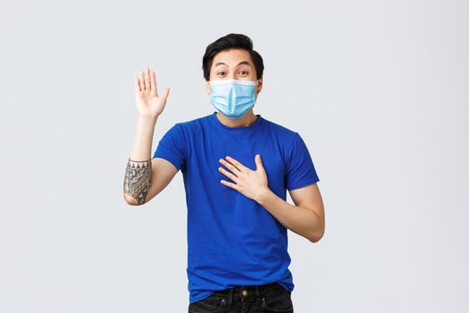 Different emotions, social distancing, self-quarantine on covid-19 and lifestyle concept. Sincere and honest asian guy in medical mask telling truth, making promise with hand raised and one on heart