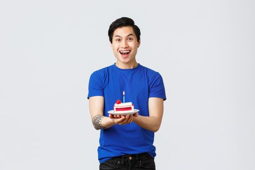 Self-quarantine, home lifestyle and celebration concept. Cheerful asian man celebrating birthday, holding piece cake and smiling upbeat, making wish, dreaming of smth, grey background.