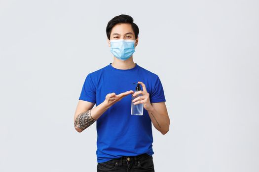 Lifestyle, people different emotions and covid-19 pandemic concept. Smiling handsome asian man in medical mask apply hand sanitizer on hand to stay safe from virus spread.