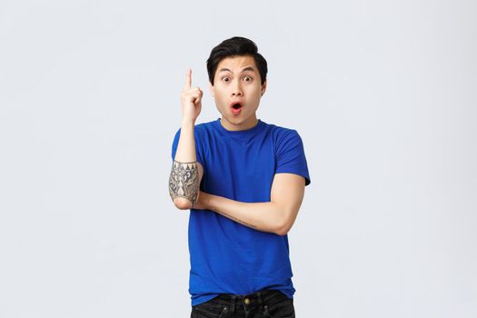 Excited handsome asian man have idea, guy in blue t-shirt raising index finger eureka sign, saying suggestion, made-up solution to problem, standing grey background