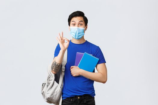 Back to school, studying during covid-19, education and university life concept. All good under control. Handsome asian male student in medical mask assure no problem with okay sign, head to class