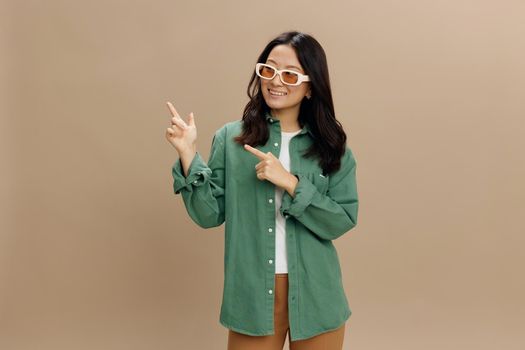Laughing happy Korean young woman in khaki green shirt stylish eyewear point fingers aside at copy space posing isolated on beige pastel studio background. Cool fashion offer. Sunglasses ad concept