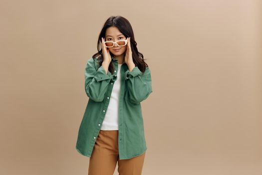 Cute Korean smiling young woman in khaki green shirt stylish touches eyewear posing isolated on over beige pastel studio background. Cool fashion offer. Sunglasses ad concept