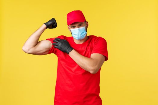 Express delivery during pandemic, covid-19, safe shipping, online shopping concept. Strong courier in red uniform, medical mask and gloves, flex biceps, show-off strong muscles