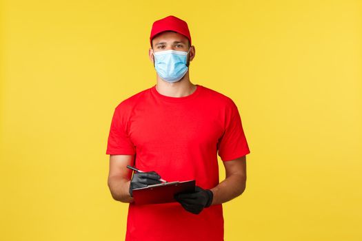 Express delivery during pandemic, covid-19, safe shipping, online shopping concept. Handsome young courier in red uniform and medical mask, writing down info on clipboard, transfer freight