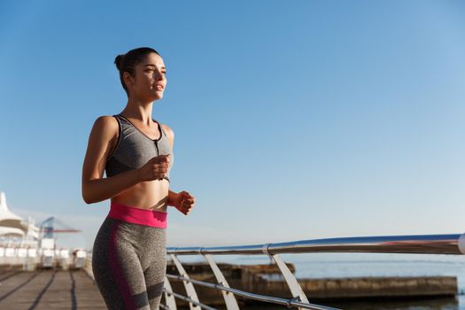 Image of young attractive sportswoman workout alone near the sea. Female athlete jogging on a pier