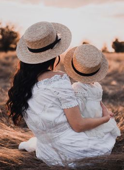 brunette mother and little daughter are sitting on the dry grass in white dresses