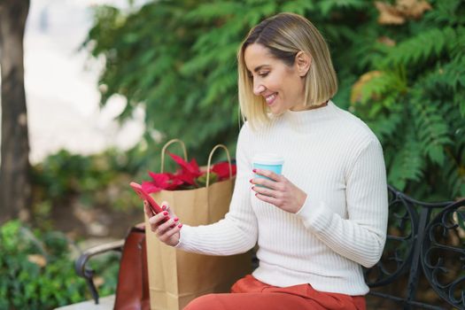 Smiling woman using smartphone and drinking beverage on bench